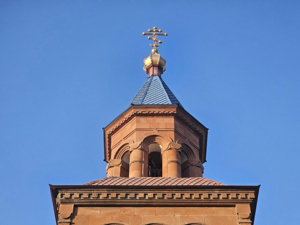 Nahaufnahme eines Kirchturms mit einem goldenen Kreuz und komplizierter Architektur vor einem klaren blauen Himmel.