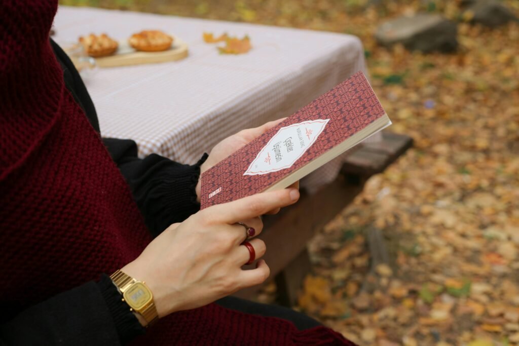 Eine Person liest im Herbst an einem Picknicktisch im Freien ein Buch und isst Gebäck. Gemütliche Herbststimmung.